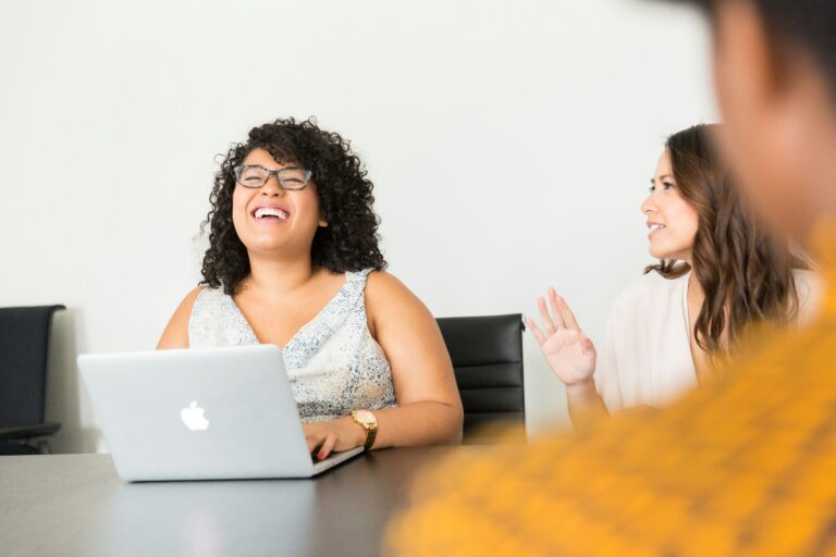 Woman at work on her laptop laughing. Woman sitting next to her talking